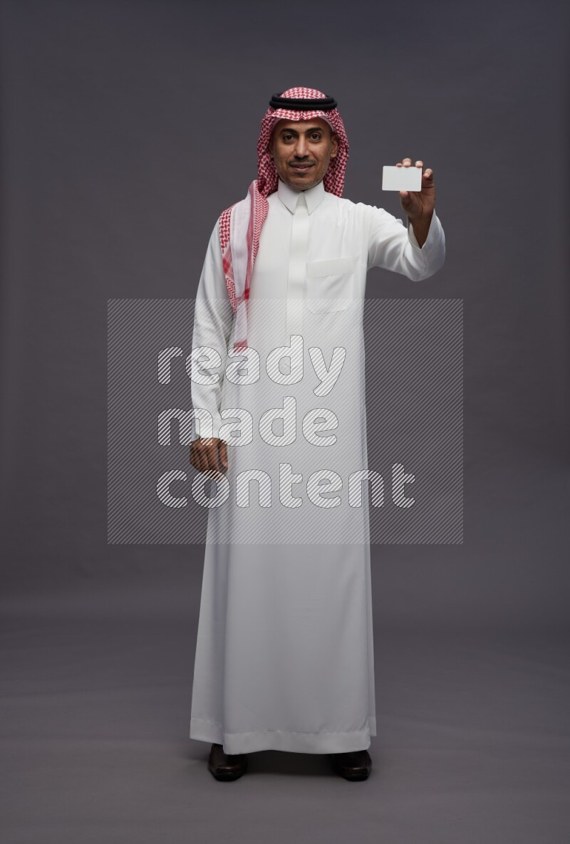 Saudi man wearing thob and shomag standing holding ATM card on gray background