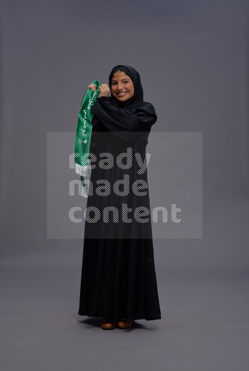 Saudi woman wearing Abaya standing holding Saudi flag on gray background