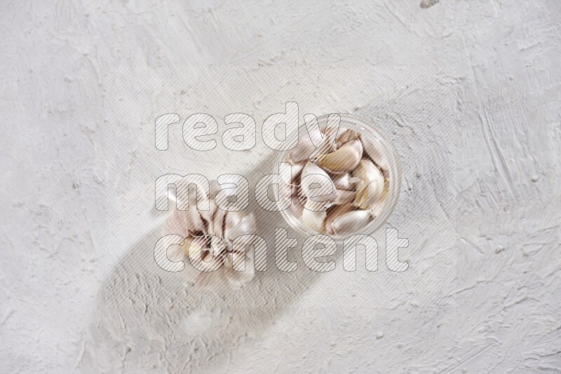 A glass jar full of garlic cloves with a whole garlic bulb beside it on a textured white flooring