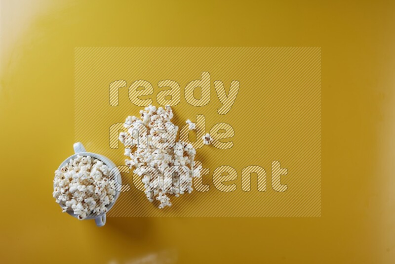 A white ceramic bowl full of popcorn with popcorn beside it on a yellow background in a top view shot