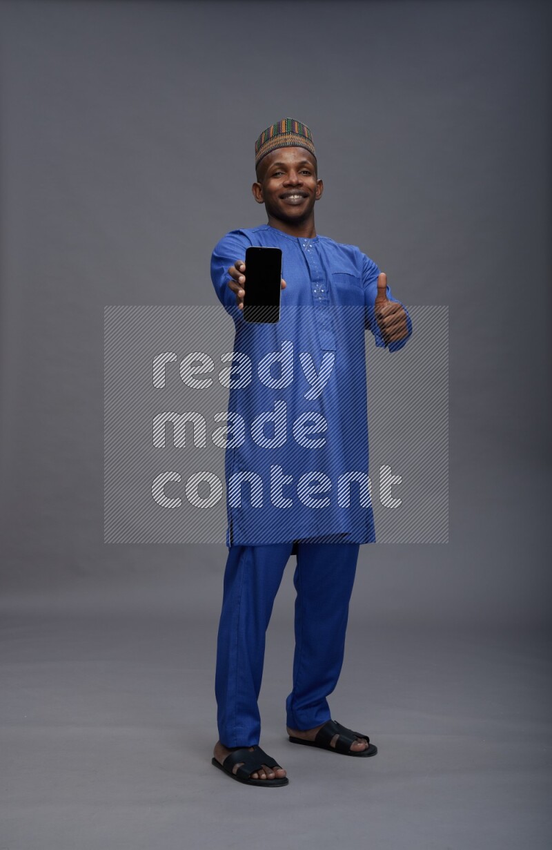 Man wearing Nigerian outfit standing showing phone to camera on gray background