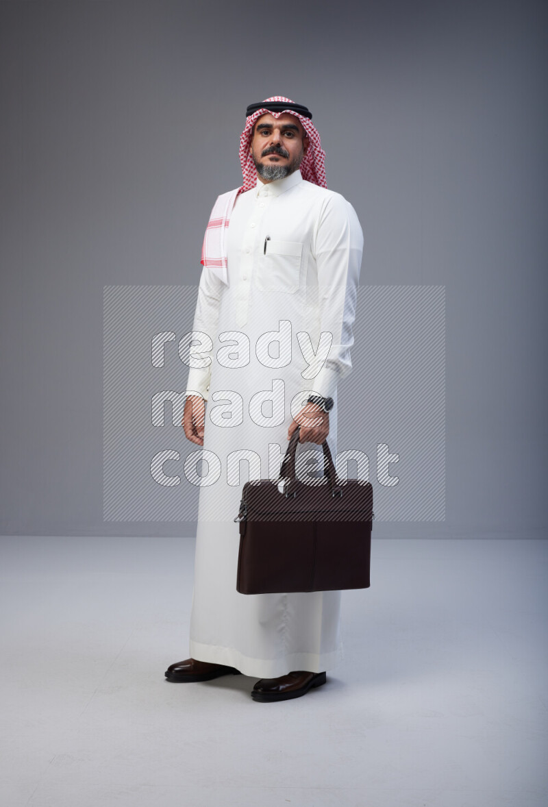 Saudi man Wearing Thob and red Shomag standing holding bag on Gray background
