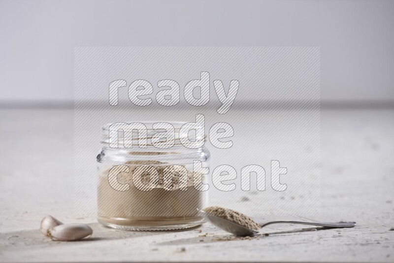 A glass jar full of garlic powder with a metal spoon full of the powder on a textured white flooring