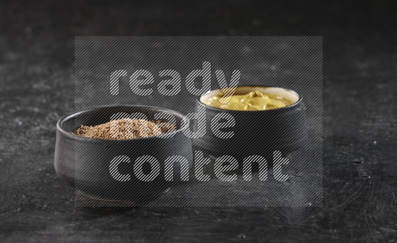 2 black pottery bowls full of mustard seeds and mustard paste on black flooring