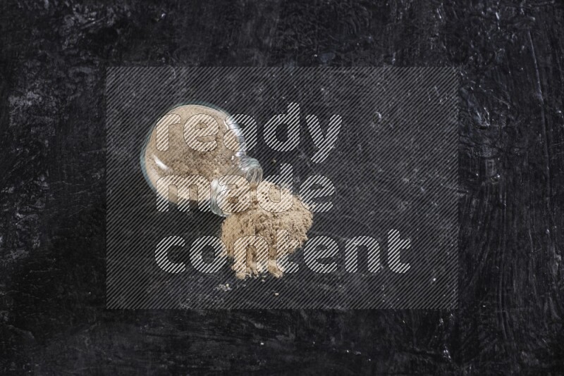 A glass spice jar full of garlic powder flipped over with the powder came out on a textured black flooring