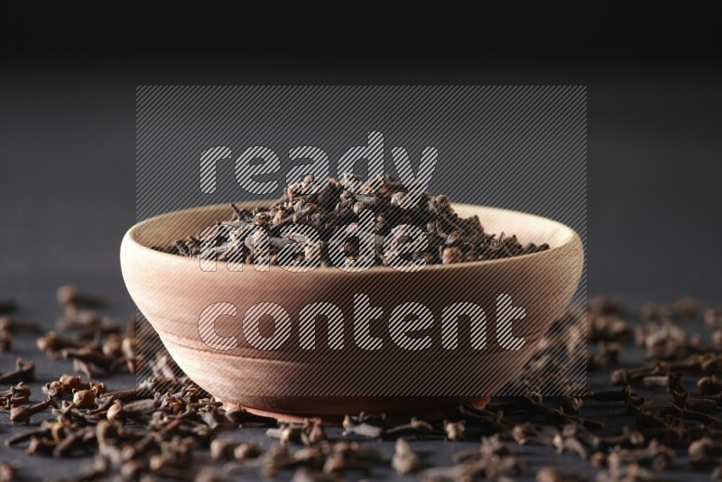 A wooden bowl full of cloves with spreaded whole cloves on a black flooring
