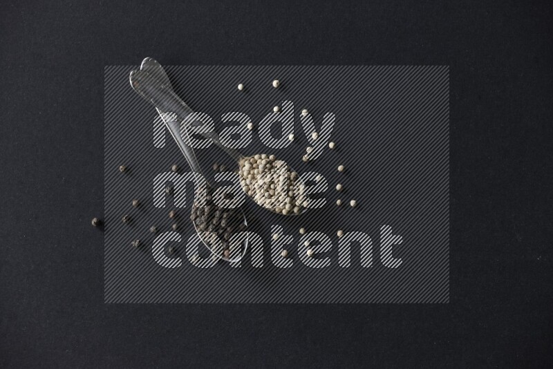 2 metal spoons full of black and white pepper beads with spreaded beads on black flooring