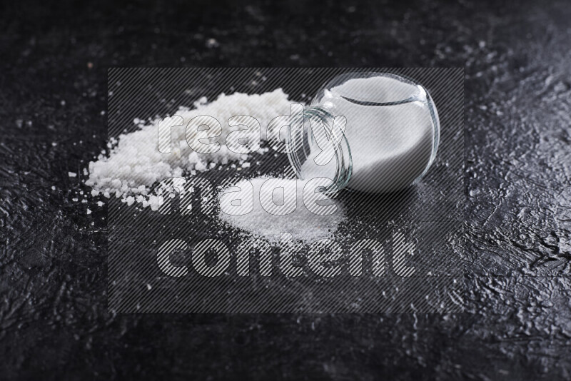 A glass jar full of table salt with some sea salt crystals beside it on a black background