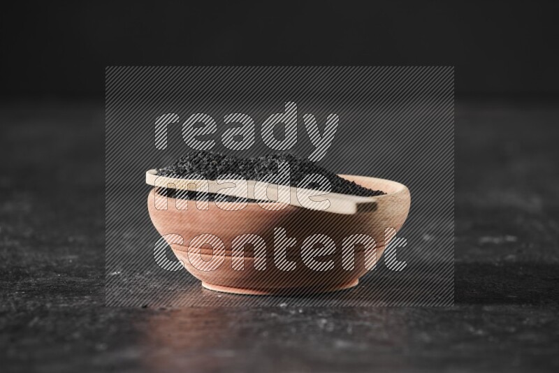 A wooden bowl full of black seeds with wooden spoon full of the seeds on it on a textured black flooring