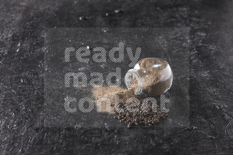 A flipped glass spice jar full of black pepper powder with pepper beads on textured black flooring