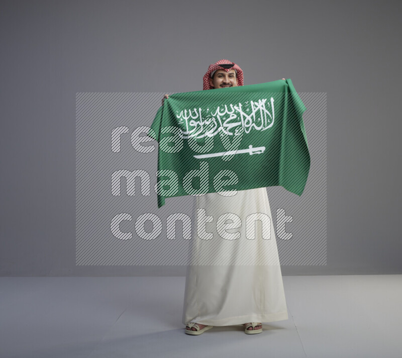 A saudi man standing wearing thob and red shomag holding big saudi flag on gray background