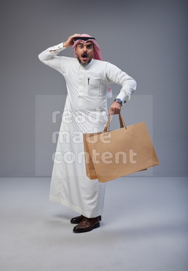 Saudi man Wearing Thob and red Shomag standing holding shopping bag on Gray background