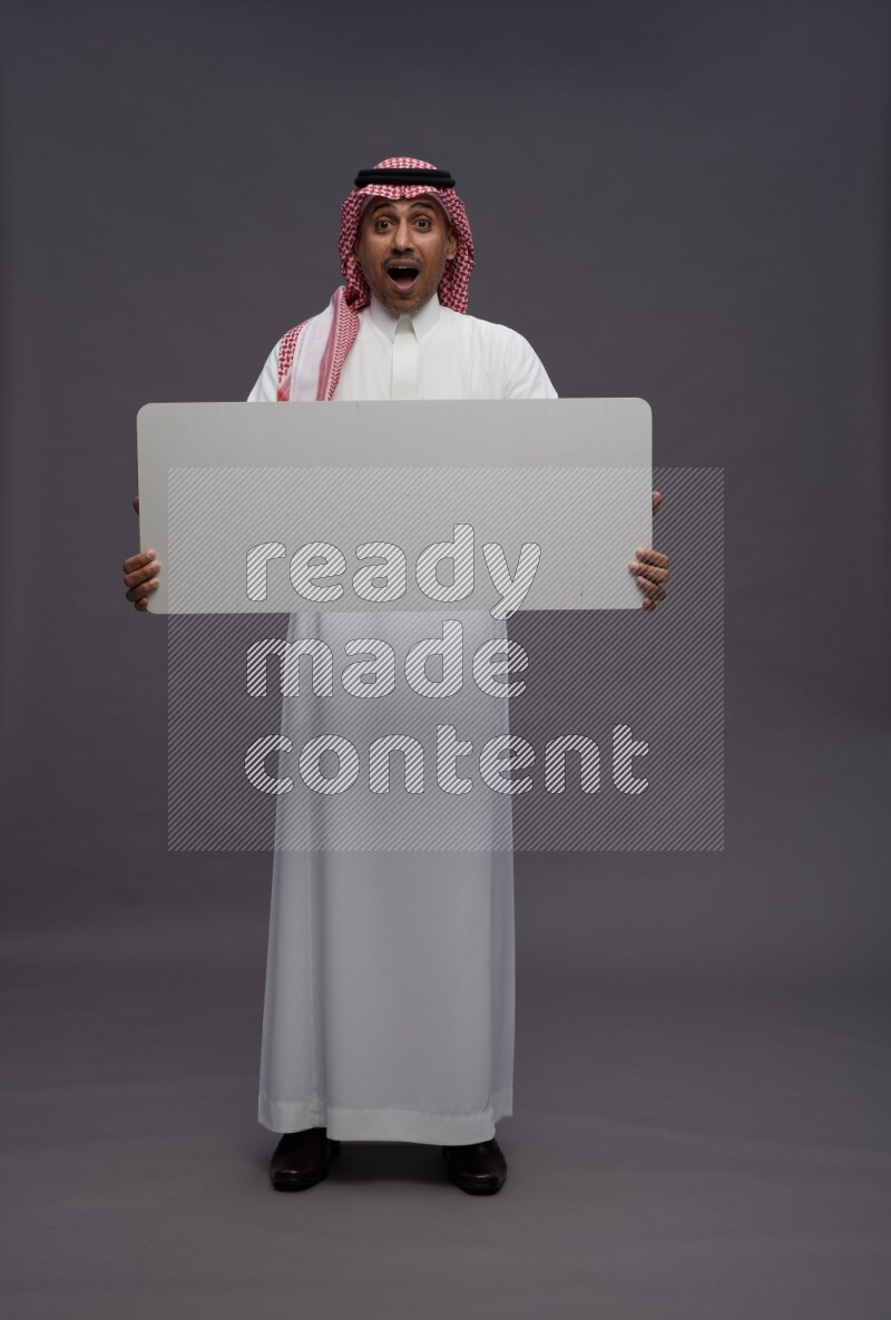 Saudi man wearing thob and shomag standing holding board on gray background