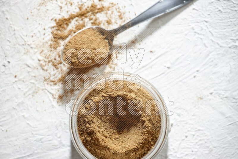 A glass jar and a metal spoon full of cumin powder on textured white flooring