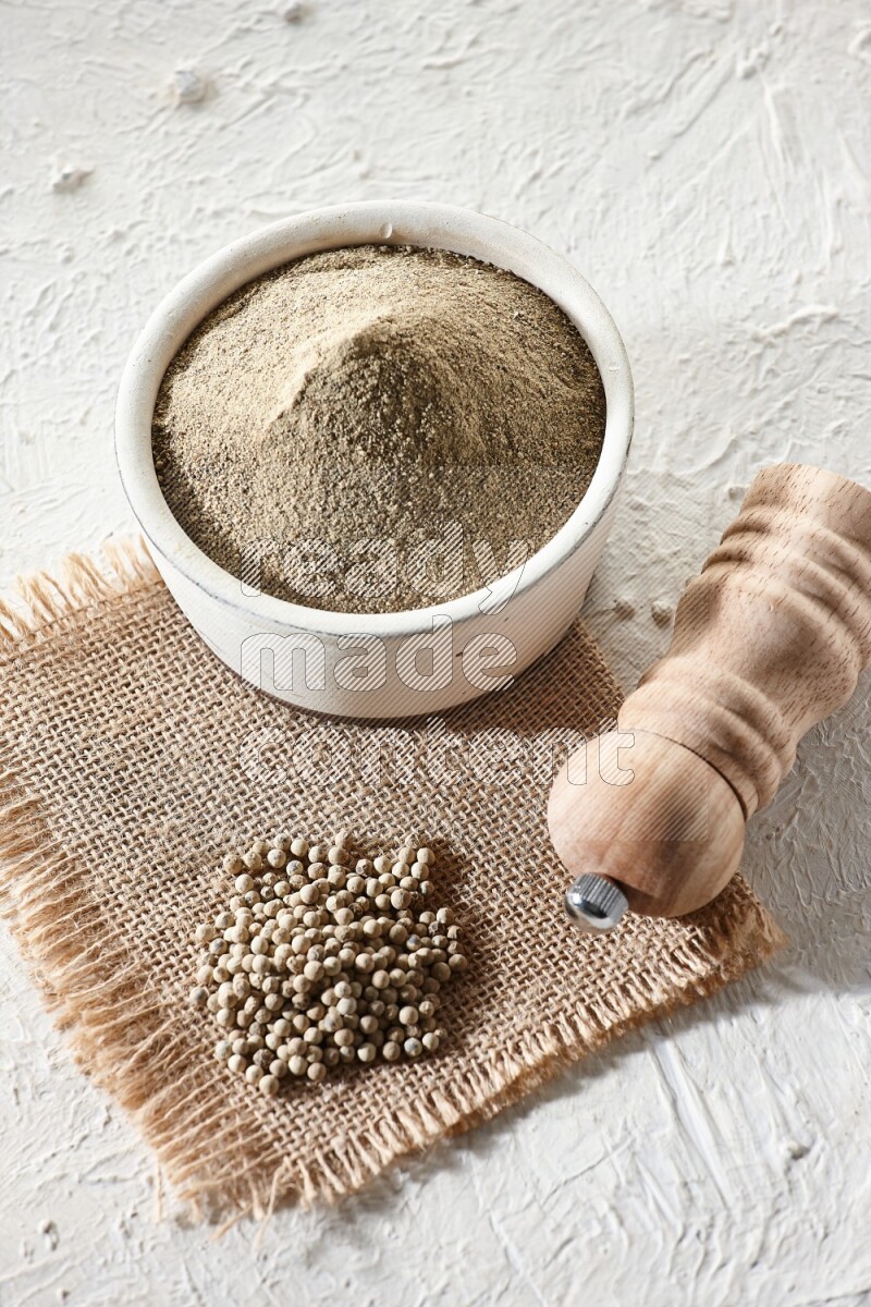 White pottery bowl full of white pepper powder set on a burlap piece of fabric with pepper beads and wooden pepper grinder on textured white flooring