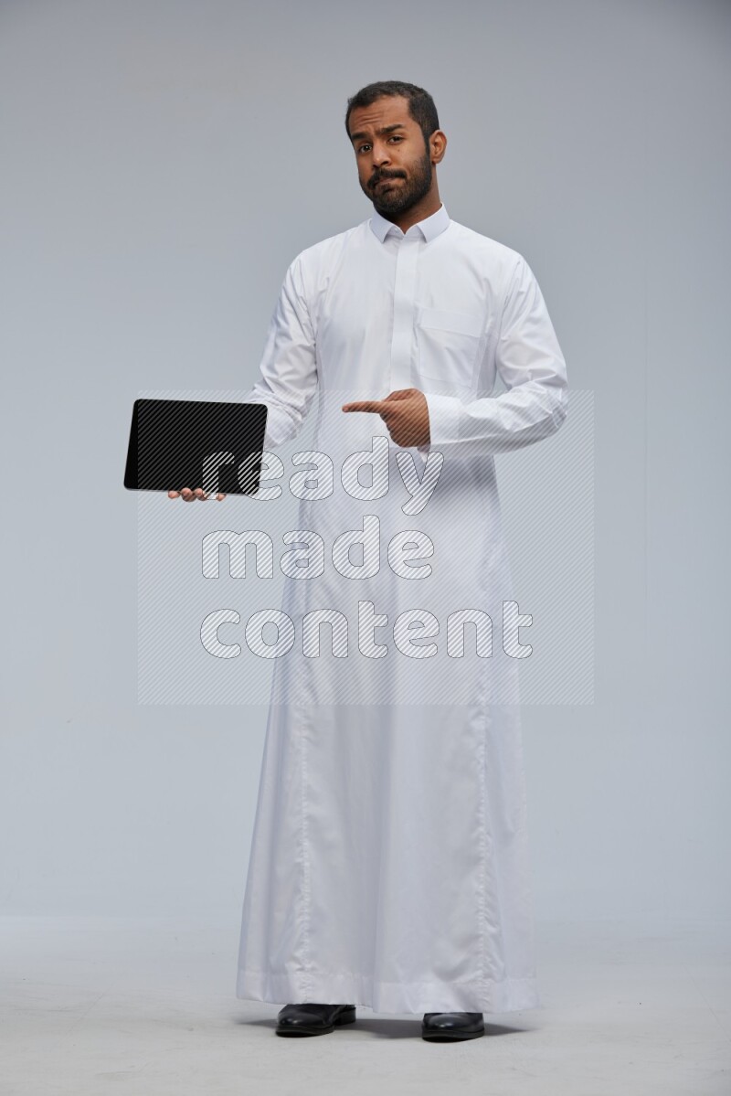 Saudi man wearing Thob standing showing tablet to camera on Gray background