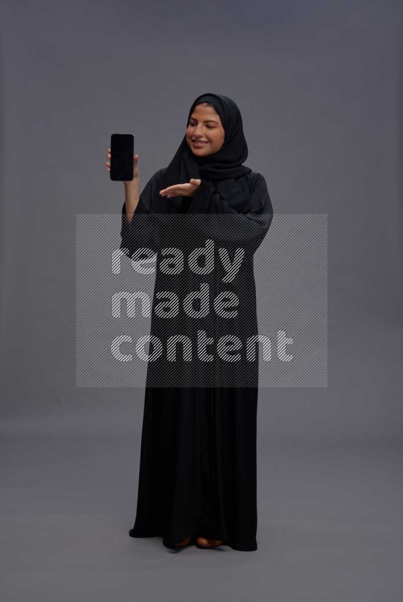 Saudi woman wearing Abaya standing showing phone to camera on gray background
