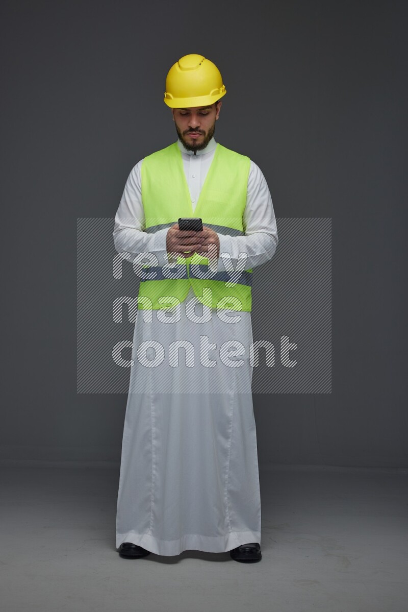 A Saudi man wearing Thobe with a yellow safety vest and white helmet standing and using his phone eye level on a gray background