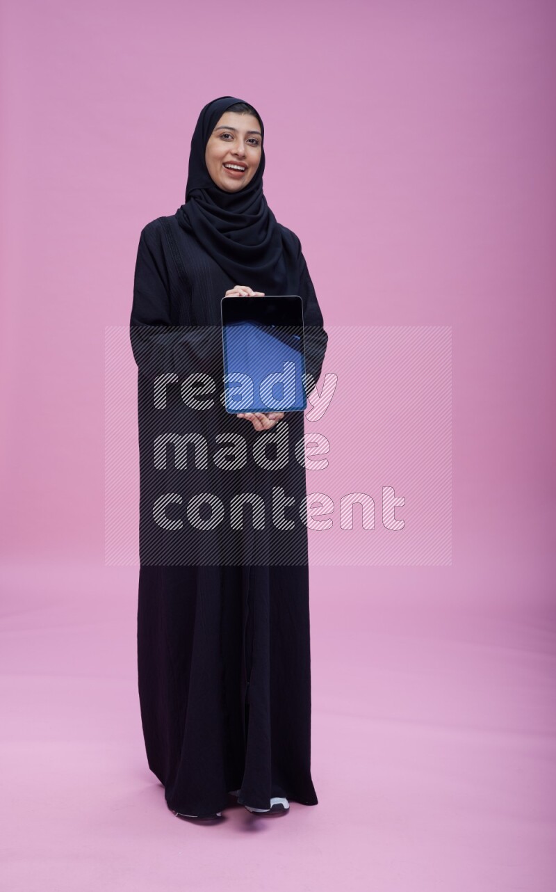 Saudi woman wearing Abaya standing showing tablet to camera on pink background
