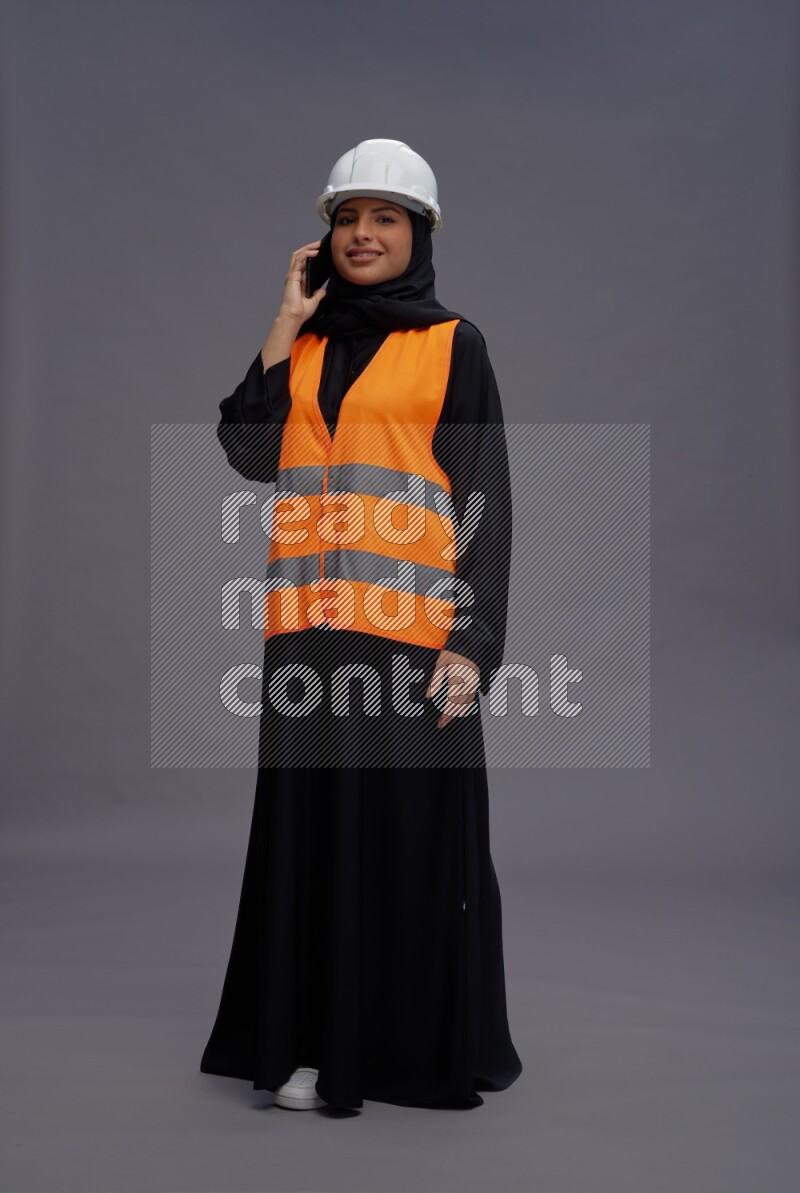 Saudi woman wearing Abaya with engineer vest standing talking on phone on gray background