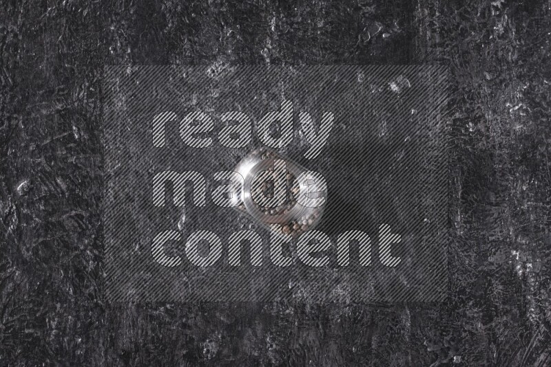 A glass spice jar full of black pepper beads on textured black flooring