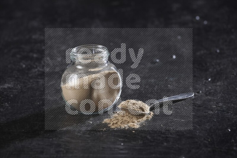 A glass spice jar full of garlic powder with a filled metal spoon on a textured black flooring