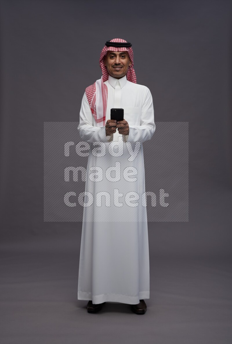 Saudi man wearing thob and shomag standing texting on phone on gray background