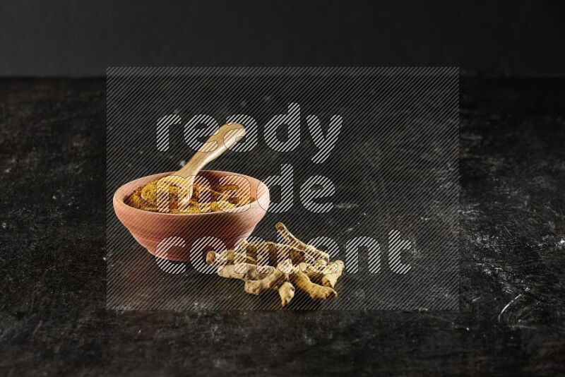 A wooden bowl and a wooden spoon full of turmeric powder with dried turmeric fingers on textured black flooring