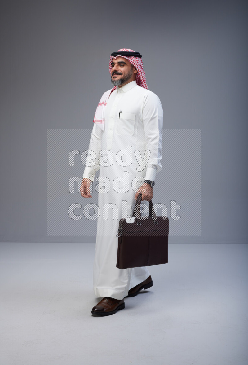 Saudi man Wearing Thob and red Shomag standing holding bag on Gray background