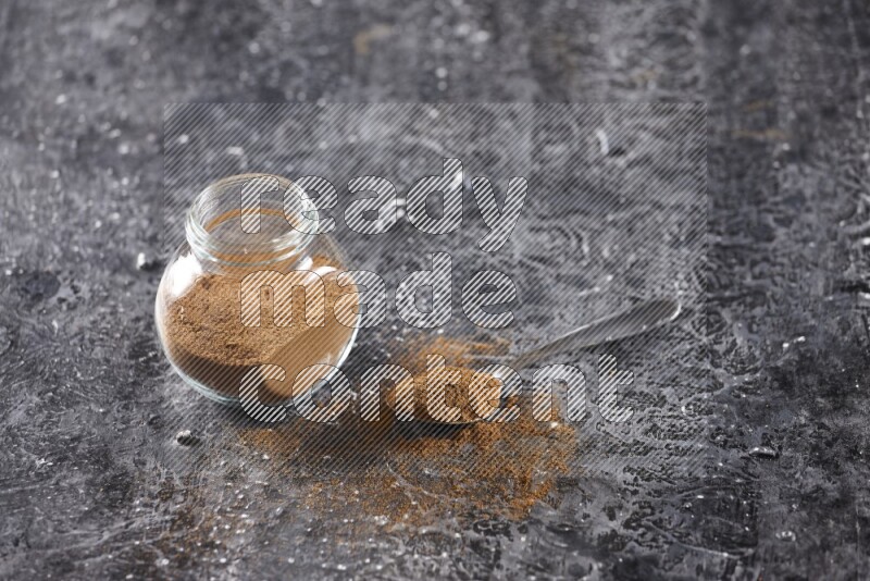 Herbal glass jar full of cinnamon powder and a metal spoon full of powder on textured black background