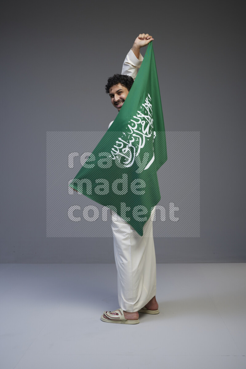 A Saudi man standing wearing thob holding big Saudi flag on gray background