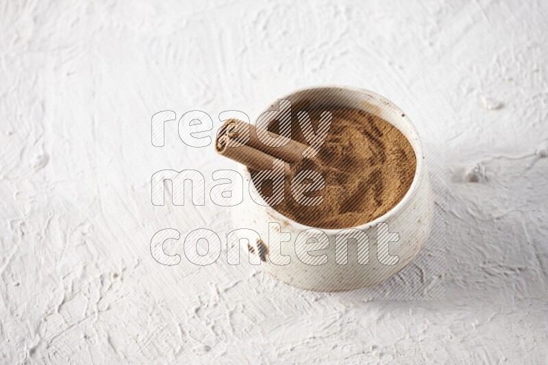 Ceramic beige bowl full of cinnamon powder with a cinnamon stick on a textured white background