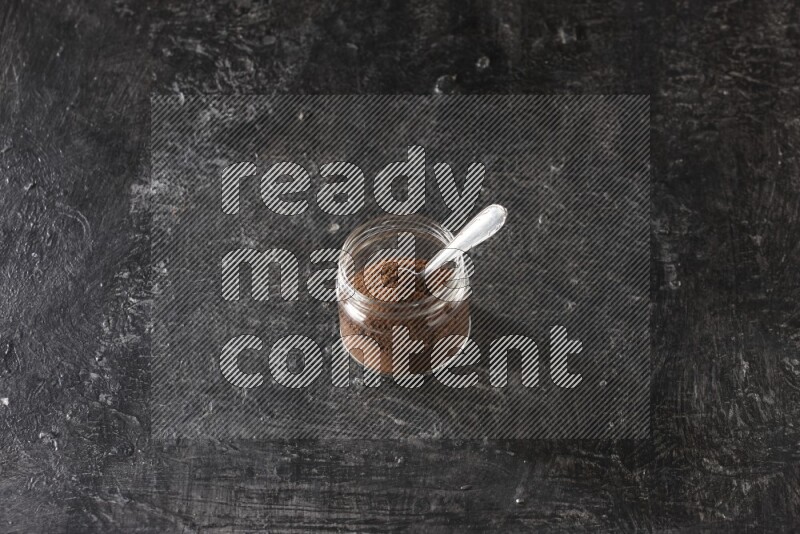 A glass jar full of cloves powder with a metal spoon on a textured black flooring