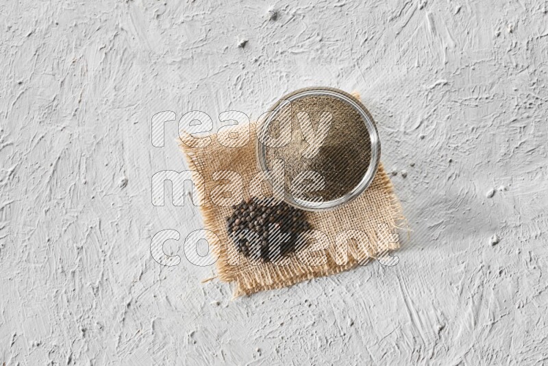 A glass bowl full of black pepper powder and black pepper beads on burlap fabric on textured white flooring