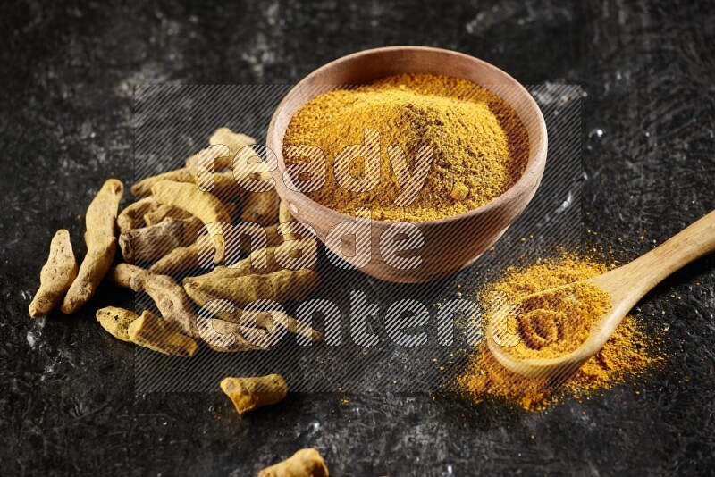 A wooden bowl and a wooden spoon full of turmeric powder with dried turmeric fingers on textured black flooring