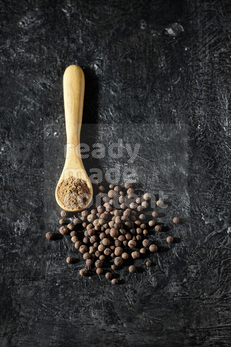 A wooden spoon full of allspice powder and whole balls spreaded on a textured black flooring