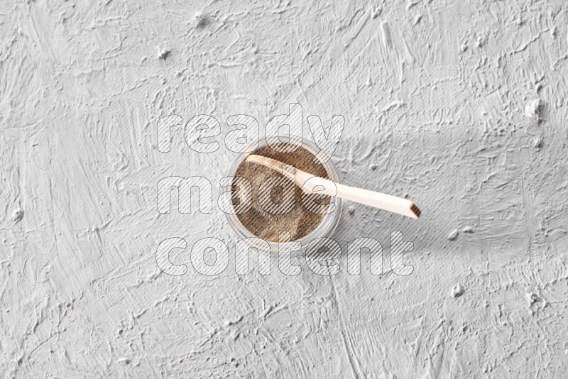 A glass jar full of black pepper powder with a wooden spoon on a textured white flooring