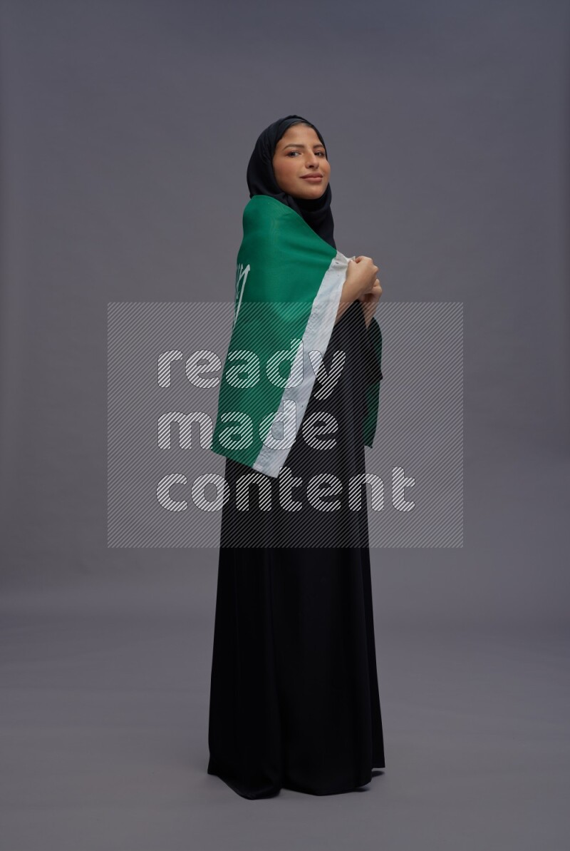 Saudi woman wearing Abaya standing holding Saudi flag on gray background