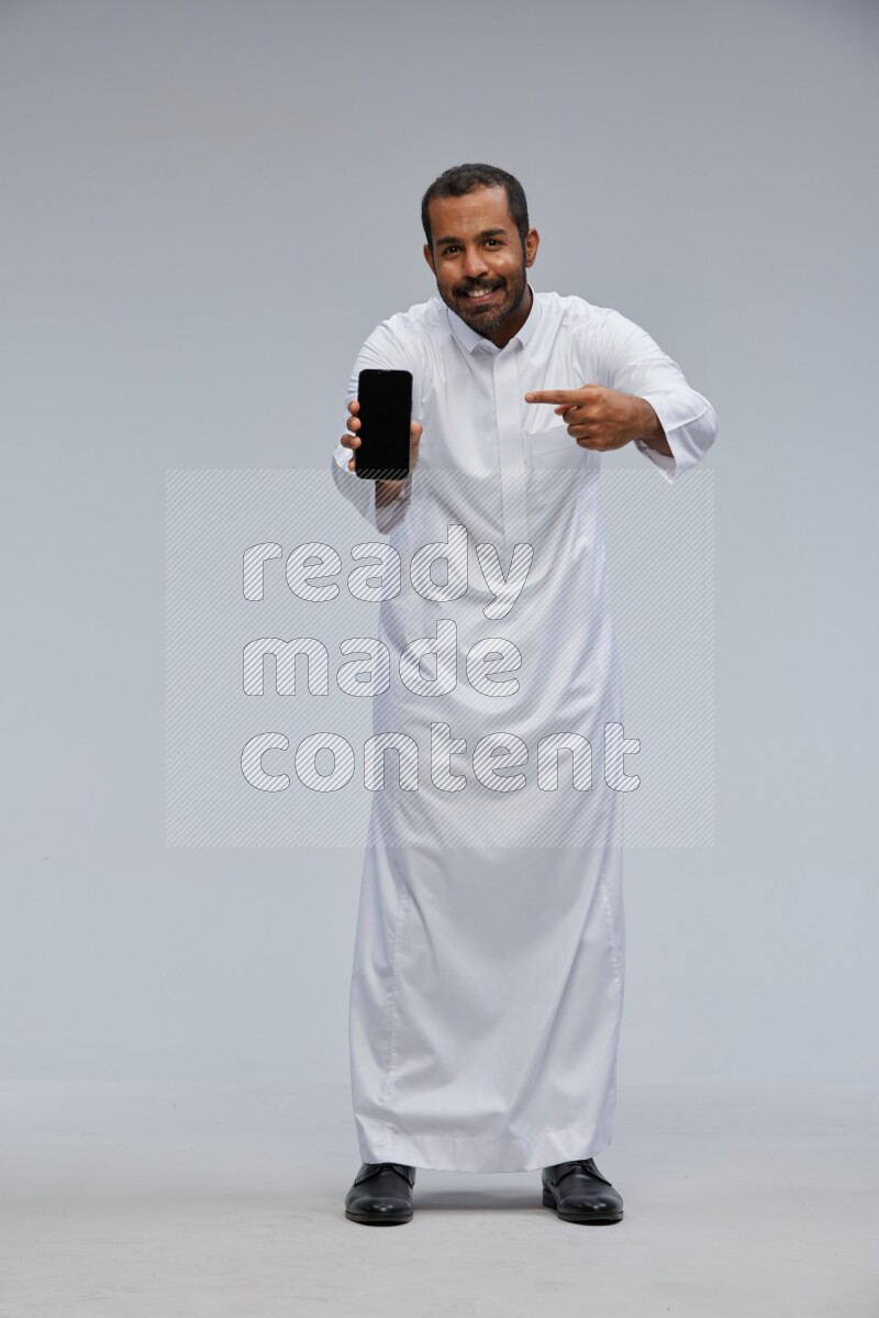 Saudi man wearing Thob standing showing phone to camera on Gray background