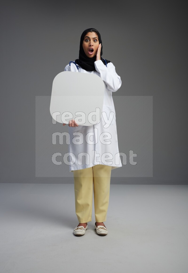 Saudi woman wearing lab coat with stethoscope standing holding social media sign on Gray background