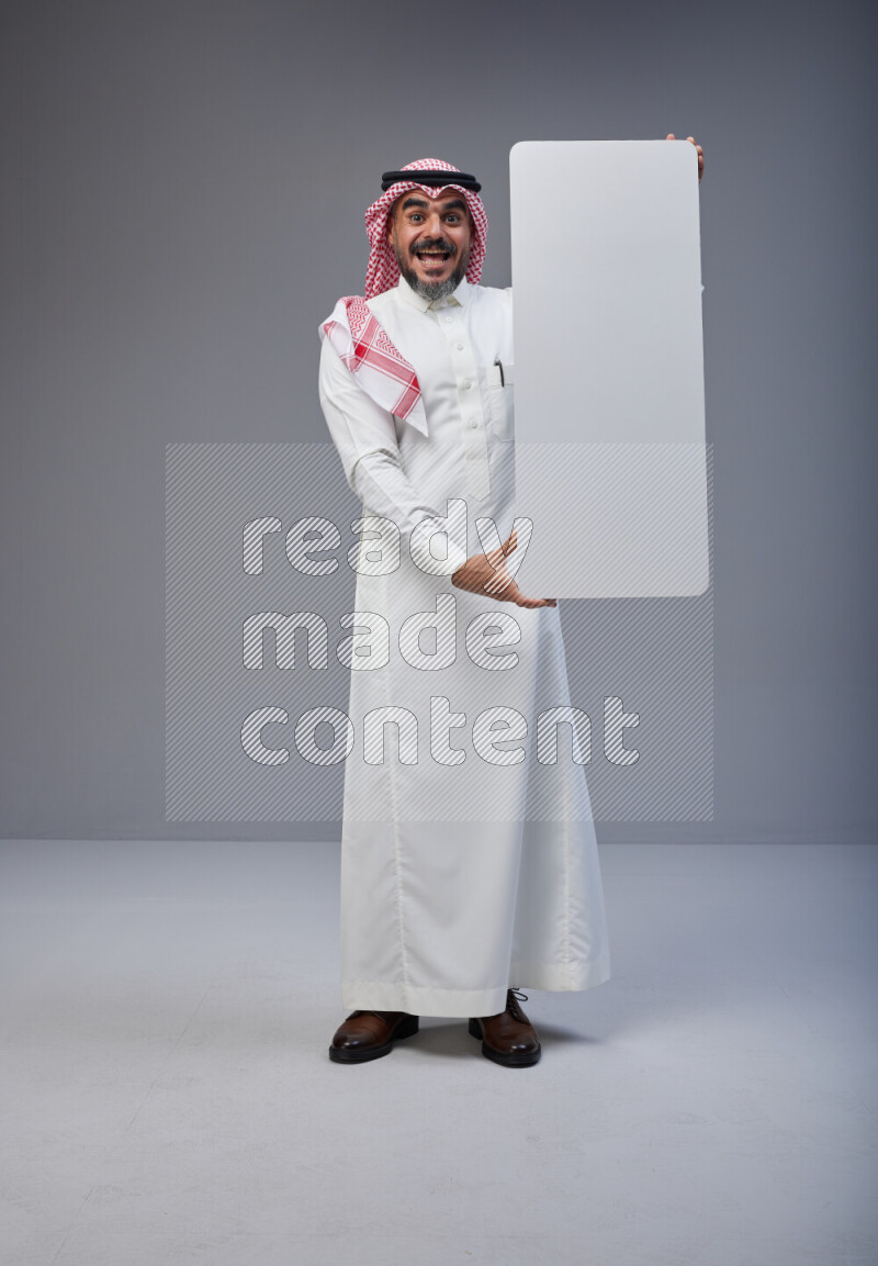 Saudi man Wearing Thob and red Shomag standing holding board on Gray background