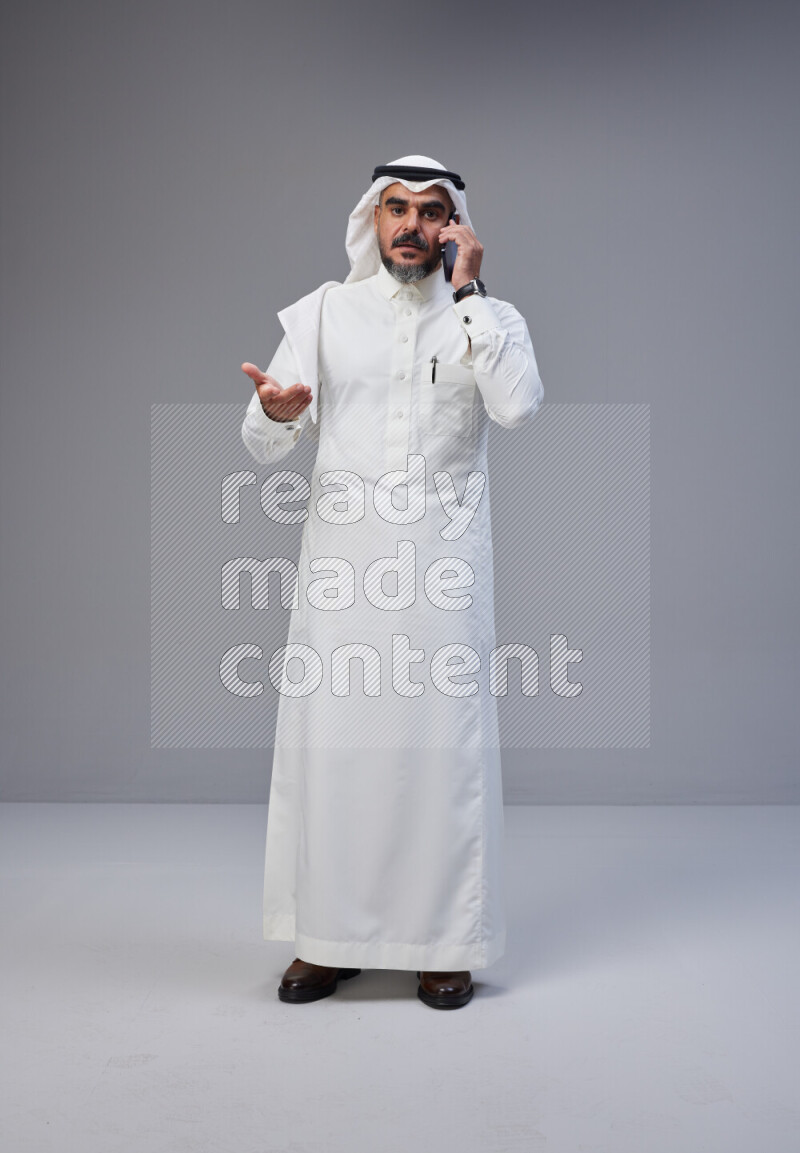 Saudi man Wearing Thob and white Shomag standing talking on phone on Gray background