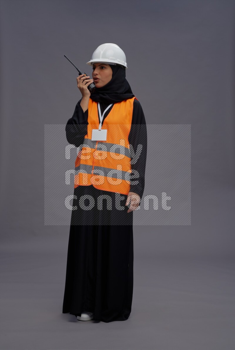 Saudi woman wearing Abaya with engineer vest with neck strap employee badge standing holding walkie-talkie on gray background