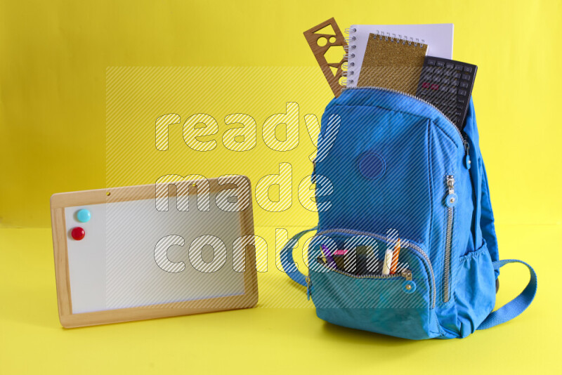 A school bag with assorted school supplies in and beside it on yellow background