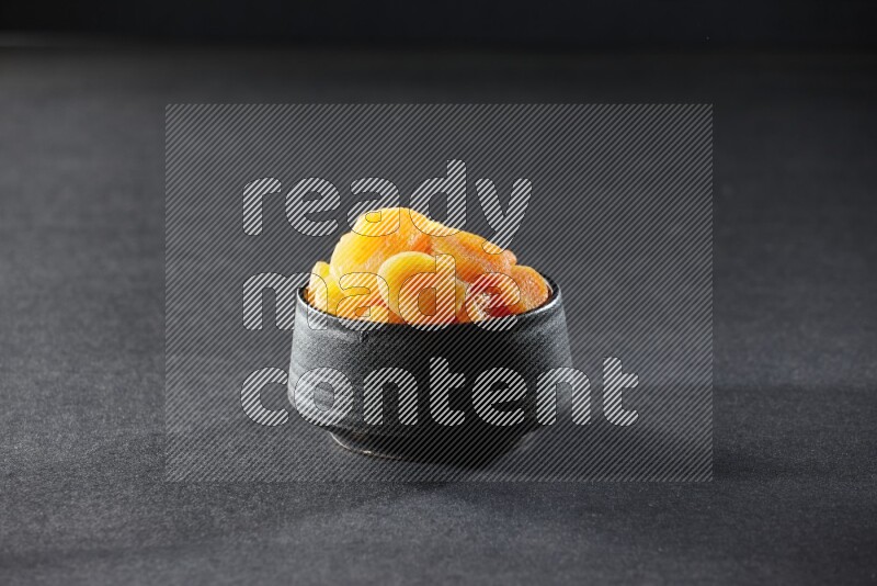 A black pottery bowl full of dried apricots on a black background in different angles