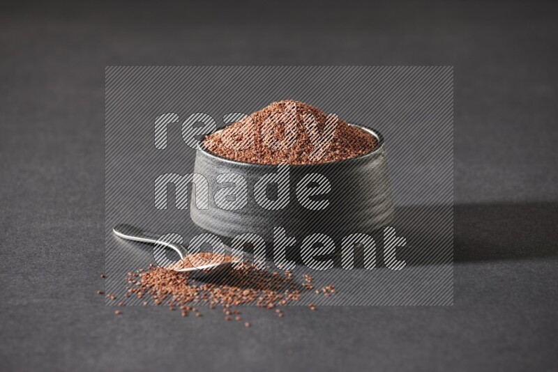 وعاء فخاري أسود ممتلئ ببذور حب الرشاد مع ملعقة معدنية ممتلئة بالبذور على أرضية سوداء