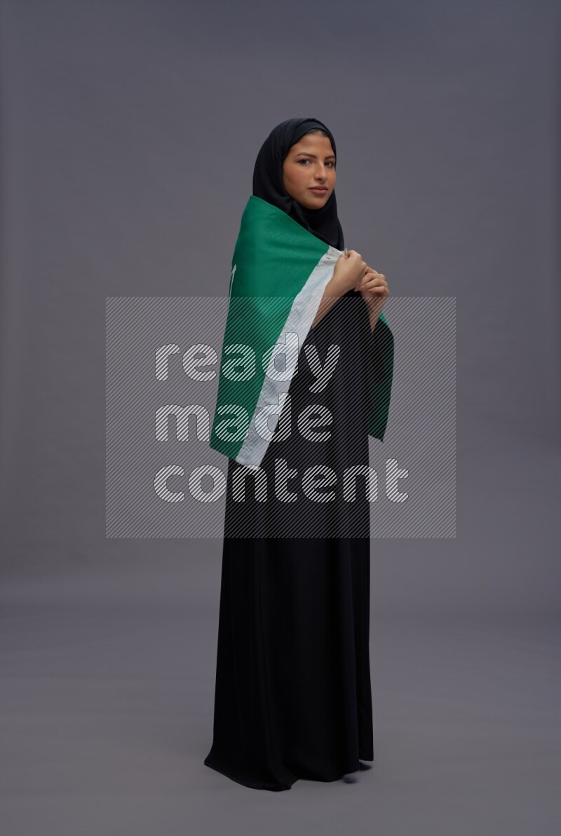 Saudi woman wearing Abaya standing holding Saudi flag on gray background
