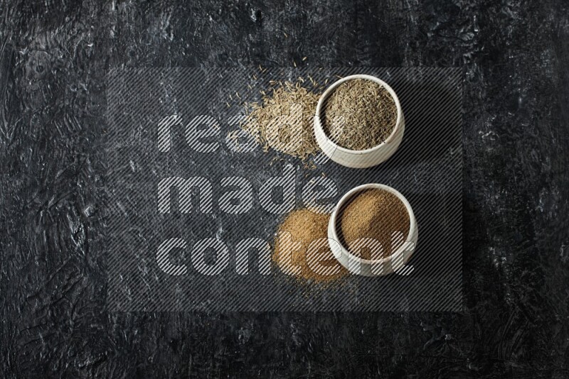 2 beige bowls, one full of cumin powder and the other full of seeds on a textured black flooring