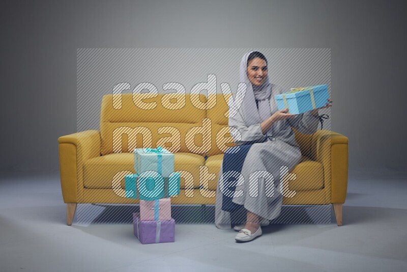 A Saudi woman wearing a light gray Abaya and head scarf sitting on a yellow sofa holding a gift box beside multi color gift boxes eye level on a grey background