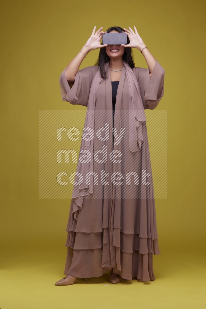 A woman Showing SmartPhone Screen on a Yellow Background wearing Brown Abaya